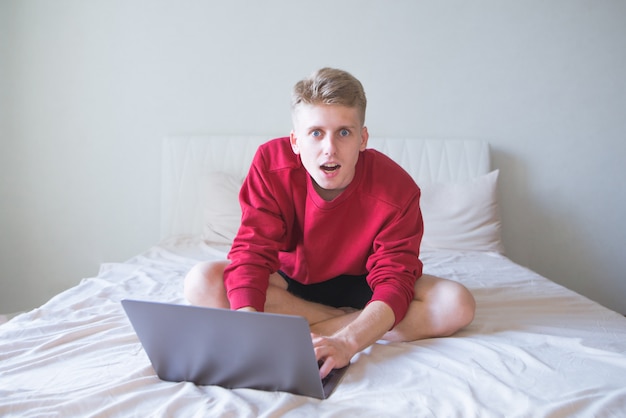 Jovem surpreendido sentado na cama em uma sala iluminada com um laptop