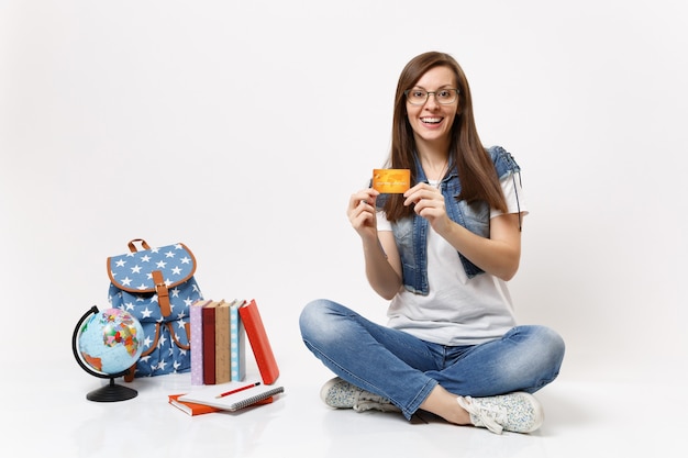 Jovem surpresa, alegre, estudante, vestindo roupas jeans de óculos, segurando um cartão de crédito, sentada perto da mochila globo, livros escolares isolados