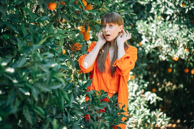 Jovem surpresa de vestido laranja está olhando para a câmera segurando a mão atrás das orelhas no jardim laranja