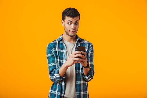 Jovem surpreso com uma camisa xadrez em pé, isolado na parede laranja, usando um telefone celular