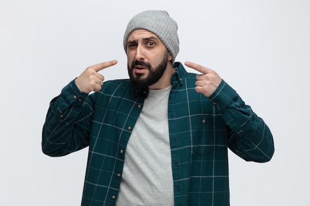 Jovem suspeito usando chapéu de inverno olhando para a câmera apontando para si mesmo isolado no fundo branco