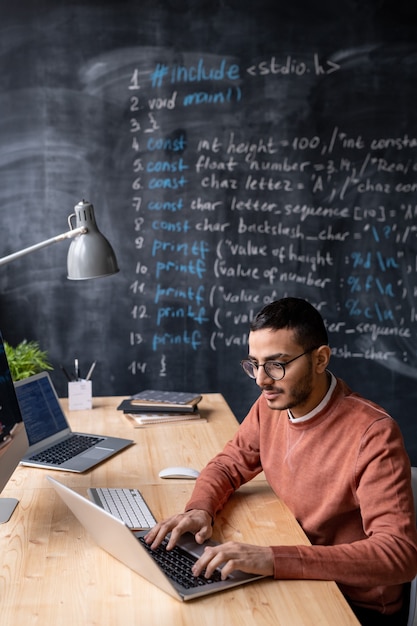 Jovem talentoso programador do Oriente Médio com barba sentado na mesa e usando o laptop enquanto trabalha no escritório com o quadro-negro