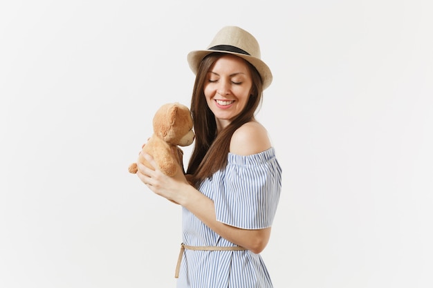 Jovem terna elegante encantadora mulher vestida de vestido azul, chapéu bonito abraço urso de pelúcia bonito brinquedo posando isolado no fundo branco. Pessoas, emoções sinceras, conceito de estilo de vida. Área de publicidade. Copie o espaço.