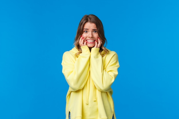 Jovem tímida e caucasiana loira caucasiana, com vergonha, com capuz amarelo em pânico, roer unhas por medo e preocupações, parecendo assustada com expressão alarmada,