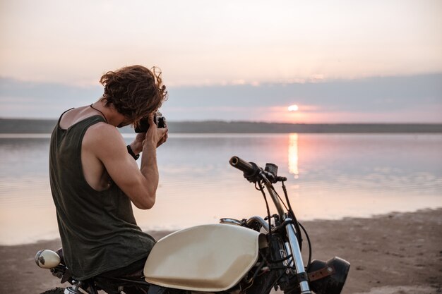 Jovem tirando fotos com a câmera enquanto está sentado em sua moto ao ar livre