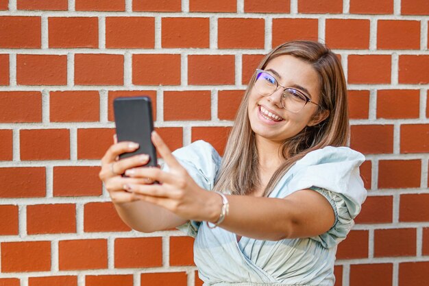 Jovem tirando uma selfie sorrindo para a câmera Estudante feliz em pé junto à parede e usando seu smartphone