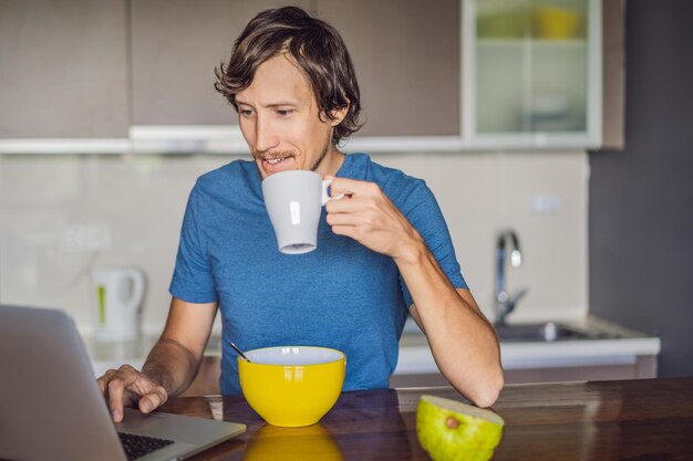 Jovem tomando café da manhã e usando laptop na cozinha