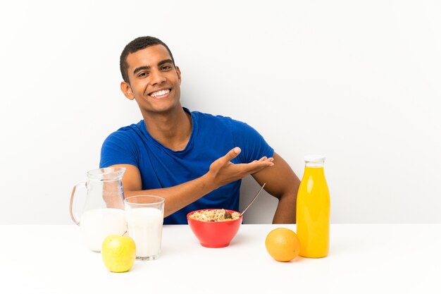 Jovem tomando café da manhã em uma mesa, estendendo as mãos para o lado para convidar para vir