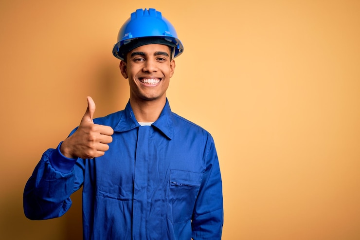 Foto jovem trabalhador afro-americano bonito vestindo uniforme azul e capacete de segurança fazendo um gesto feliz de polegar para cima com a mão, com expressão de aprovação olhando para a câmera mostrando sucesso