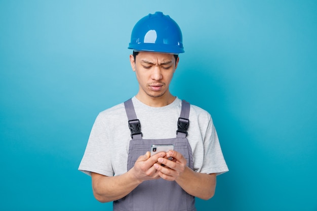Jovem trabalhador da construção civil confuso usando capacete de segurança e uniforme, segurando e olhando para o celular