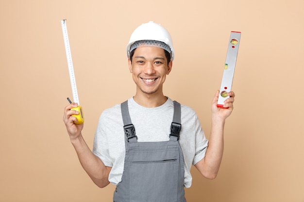 Jovem trabalhador da construção civil sorridente usando capacete de segurança e uniforme segurando régua de nível e medidor de fita adesiva