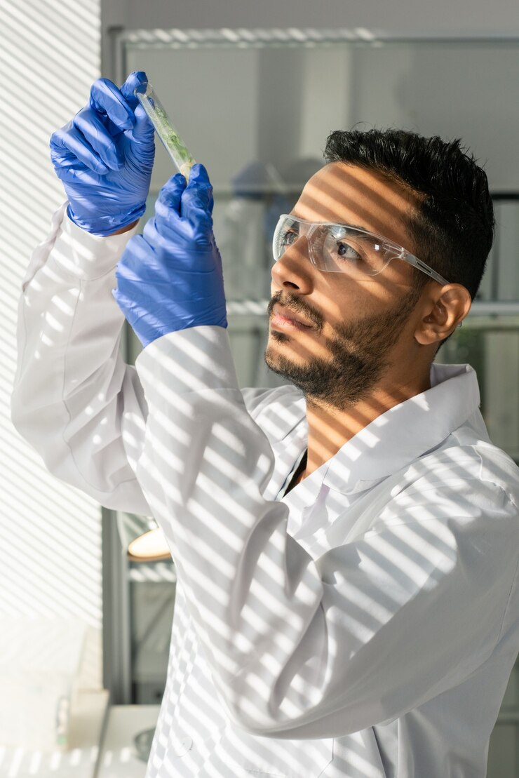 Foto jovem trabalhador de laboratório com jaleco branco, luvas e óculos de proteção olhando para um frasco contendo broto verde crescido em laboratório