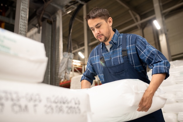 Jovem trabalhador de uma fábrica de produção de polímeros contemporânea carregando sacos com grânulos de plástico antes de processar