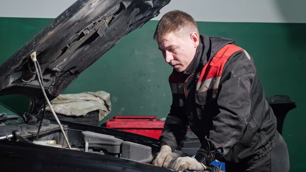 Jovem trabalhador de uniforme examina para consertar automóvel quebrado