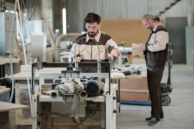 Jovem trabalhador processando peça em máquina industrial enquanto seu colega sênior ou mestre escolhe uma placa de madeira