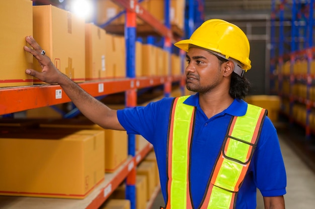 Jovem trabalhador usando capacete verificando estoque e contando produto na prateleira no armazém moderno