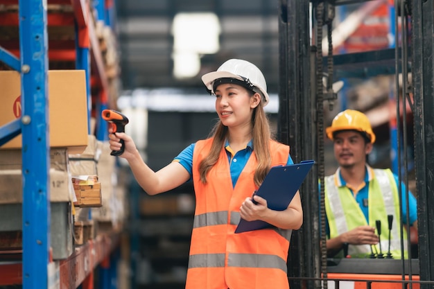 Jovem trabalhadora asiática usando scanner de código de barras verificando mercadorias e estoque de prateleira de caixa na loja de fábrica do armazém e sorria colega de trabalho feliz conduz empilhadeira nas costas conceito logístico de armazém