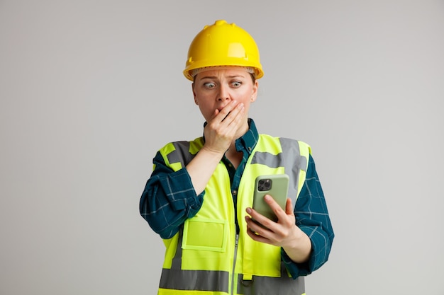 Jovem trabalhadora da construção civil preocupada usando capacete e colete de segurança, segurando e olhando para o telefone celular, mantendo a mão na boca