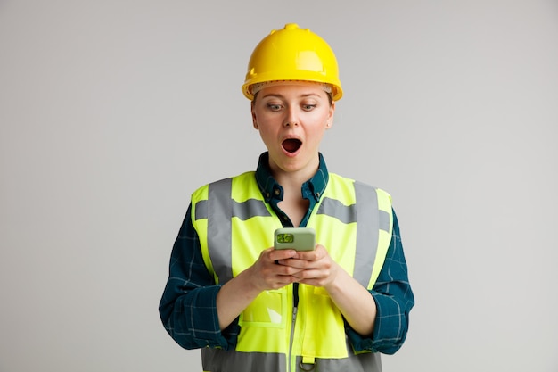 Jovem trabalhadora da construção civil surpresa usando capacete de segurança e colete de segurança segurando e olhando para o celular