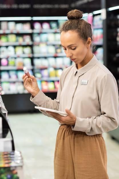 Jovem trabalhadora de um grande supermercado de beleza contemporâneo comparando as informações no touchpad com as do tubo de brilho labial