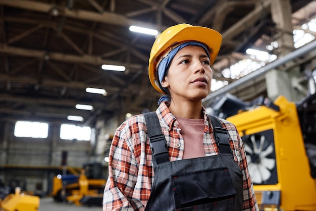 Jovem trabalhadora séria de planta industrial usando macacão e capacete de segurança