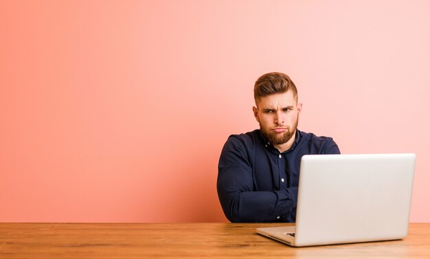 Jovem, trabalhando com seu laptop, franzindo a testa rosto em descontentamento, mantém os braços cruzados.