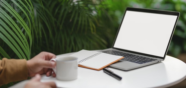 Jovem, trabalhando em seu projeto com o computador portátil enquanto bebe uma xícara de café no local de trabalho confortável ao ar livre