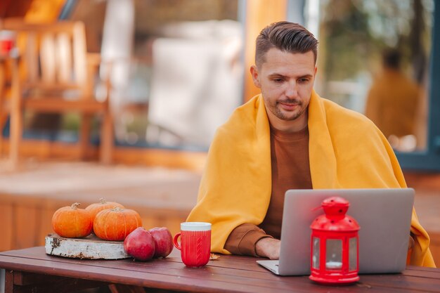 Jovem trabalhando em um laptop e bebendo café sentado à mesa de madeira ao ar livre em dia de outono