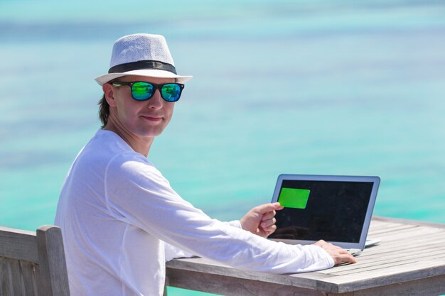 Jovem trabalhando no laptop com cartão de crédito na praia tropical