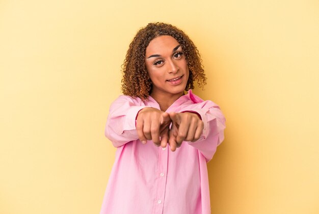 Jovem transexual latina isolada em fundo amarelo, apontando para a frente com os dedos.