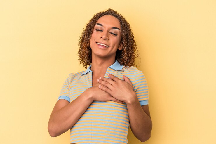 Foto jovem transexual latina isolada em fundo amarelo rindo mantendo as mãos no coração, conceito de felicidade.
