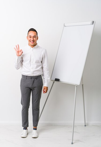 Jovem treinador latino com um quadro branco isolado pisca um olho e segura um gesto de ok com a mão.