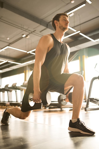 Jovem treinando no clube esportivo com halteres ele fazendo estocada e concentrando-se em seus exercícios