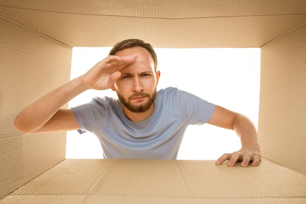 Foto jovem triste abrindo o maior pacote postal isolado no branco. modelo masculino desapontado em cima de caixa de papelão