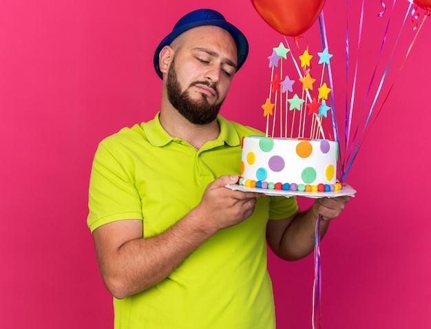 Jovem triste com chapéu de festa azul segurando balões e olhando para um bolo na mão