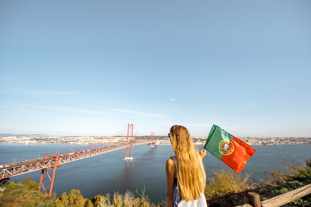 Jovem turista apreciando a bela vista aérea da famosa ponte de ferro sentado com a bandeira portuguesa na cidade de Lisboa, Portugal