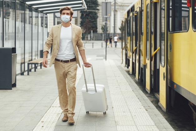 Jovem turista do sexo masculino vestido com roupa formal e máscara médica carregando bagagem na parada do bonde