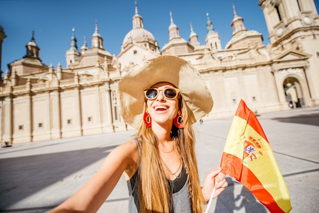 Foto jovem turista em pé com a bandeira espanhola em frente à famosa catedral na praça central durante o tempo ensolarado na cidade de zaragoza, espanha