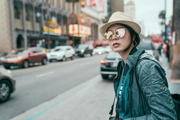 Jovem turista linda mulher asiática em óculos de sol e chapéu ao ar livre na cidade esperando táxi de táxi. conceito de viajante de transporte de viagens. garota parada na estrada na calçada da fama de hollywood boulevard.