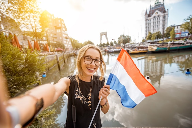 Jovem turista tirando fotos de selfie com a bandeira holandesa parada no antigo porto da cidade de Rotterdam