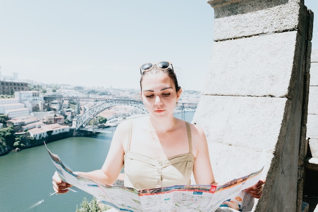 Jovem turista usando óculos escuros, olhando um mapa no meio de uma rua e sorrindo, pensando para a câmera, dia de verão, ensolarado, porto, cidade mediterrânea,