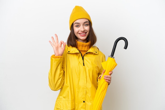 Jovem ucraniana com casaco à prova de chuva e guarda-chuva isolado no fundo branco, mostrando sinal de ok com os dedos