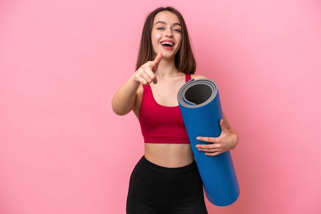 Jovem ucraniana esportiva indo para aulas de ioga enquanto segura um tapete isolado no fundo rosa surpreso e apontando para a frente