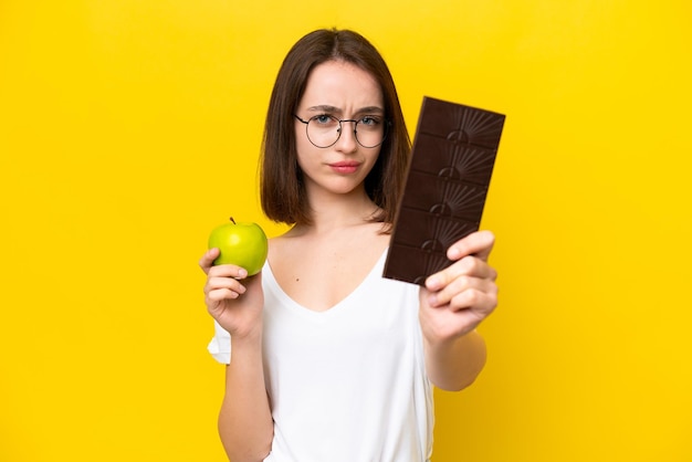 Jovem ucraniana isolada em fundo amarelo tomando um tablete de chocolate em uma mão e uma maçã na outra