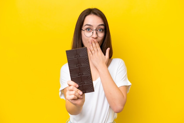 Jovem ucraniana isolada em fundo amarelo tomando uma tablete de chocolate e surpresa