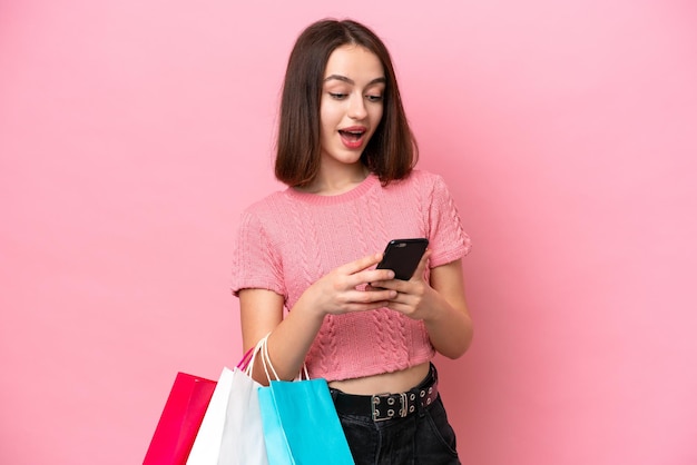 Foto jovem ucraniana isolada em fundo rosa segurando sacolas de compras e escrevendo uma mensagem com seu celular para um amigo