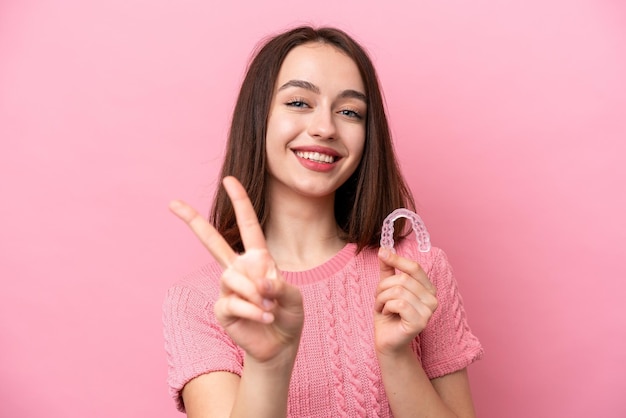 Jovem ucraniana segurando aparelho invisível isolado em fundo rosa sorrindo e mostrando sinal de vitória