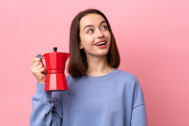 Jovem ucraniana segurando cafeteira isolada em fundo rosa olhando para cima enquanto sorria