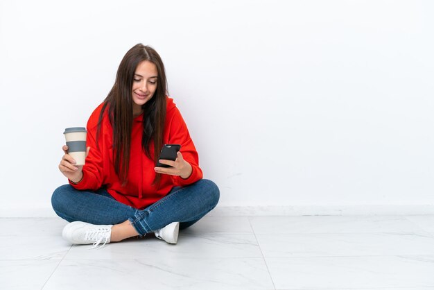 Jovem ucraniana sentada no chão isolada em fundo branco segurando café para levar e um celular