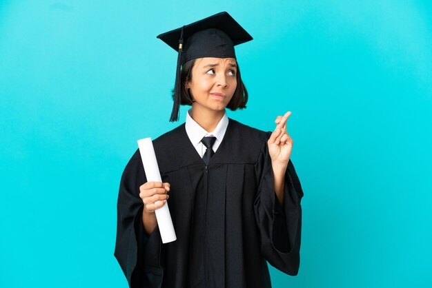 Jovem universitária sobre um fundo azul isolado com os dedos se cruzando e desejando o melhor